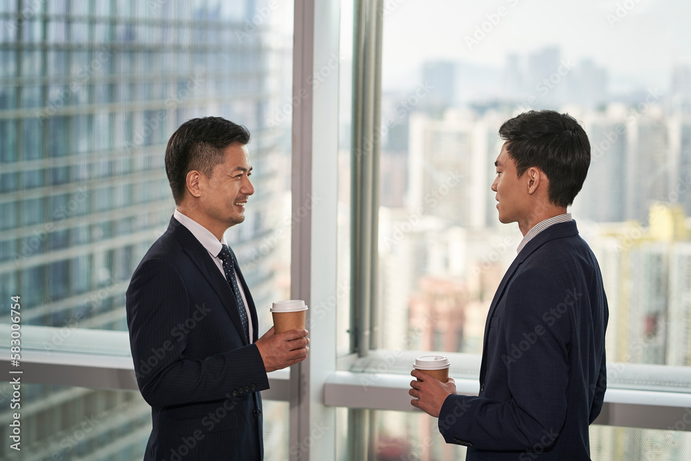 two asian business men standing and talking by the window in office two asian business men standing and talking by the window in office
