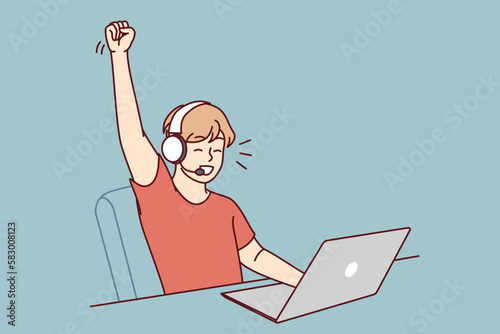 Delighted teen boy sitting at table with laptop playing video game and defeating virtual opponents. Schoolboy in headphones makes victory gesture overjoyed at broken record in internet game 
