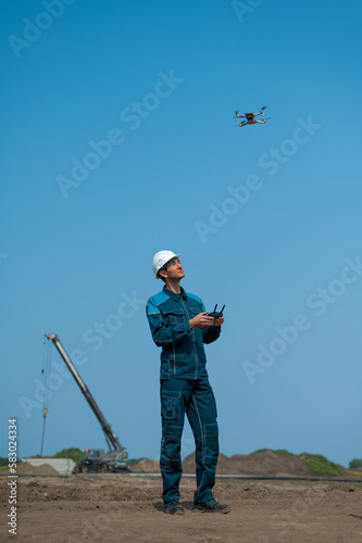 A man in a helmet and overalls controls a drone at a construction site. The builder carries out technical oversight.