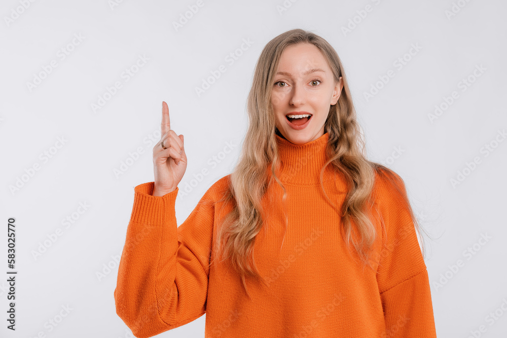 Smiling attractive young woman points finger up, shows way to banner, demonstrates logo or advertisement on top, stands in an soft orange sweater over white background Smiling attractive young woman points finger up, shows way to banner, demonstrates logo or advertisement on top, stands in an soft orange sweater over white background