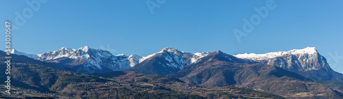 A scenic panoramic view of majestic snowy mountain range with pine forest under a majestic blue sky