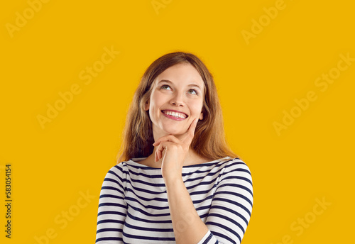 Happy woman dreaming or thinking about something nice. Beautiful young girl standing isolated on yellow background, holding hand on chin, looking up with happy, dreamy face expression and smiling