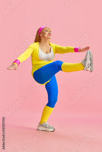 Sportive middle-aged, mature woman in colorful sportswear training, rising legs to hands, posing against pink studio background. Concept of sportive lifestyle, retirement, health care, wellness. Ad