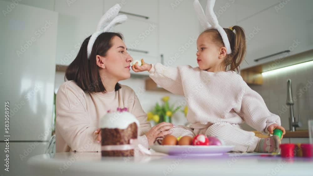 A cute daughter feeds her mother with an egg against the background of a white kitchen. Happy Easter. A cute daughter feeds her mother with an egg against the background of a white kitchen. Happy Easter.