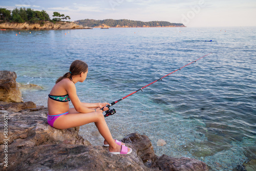 Girl fishing on the sea at rocky shore in beautiful sunset light. Summer travel holiday. 