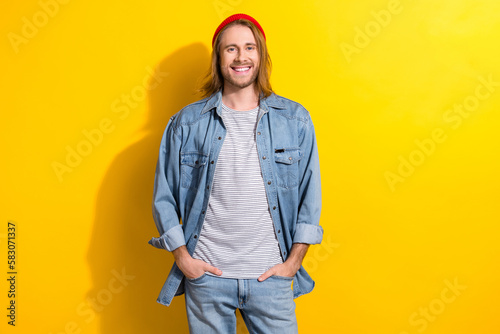 Photo of friendly handsome guy with long hairdo dressed jeans shirt standing holds arms in pockets isolated on yellow color background © deagreez