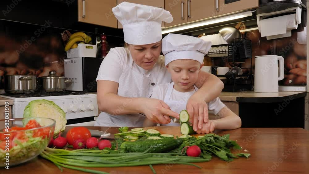 Close-up of a small child himself cutting a fresh cucumber with a knife on a wooden cutting board. Mother teaches son to cook. The concept of family relations. Close-up of a small child himself cutting a fresh cucumber with a knife on a wooden cutting board. Mother teaches son to cook. The concept of family relations.