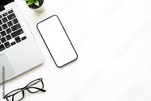 Smartphone with blank empty mockup screen is on top of white office desk table with laptop computer and supplies. Top view with copy space, flat lay.