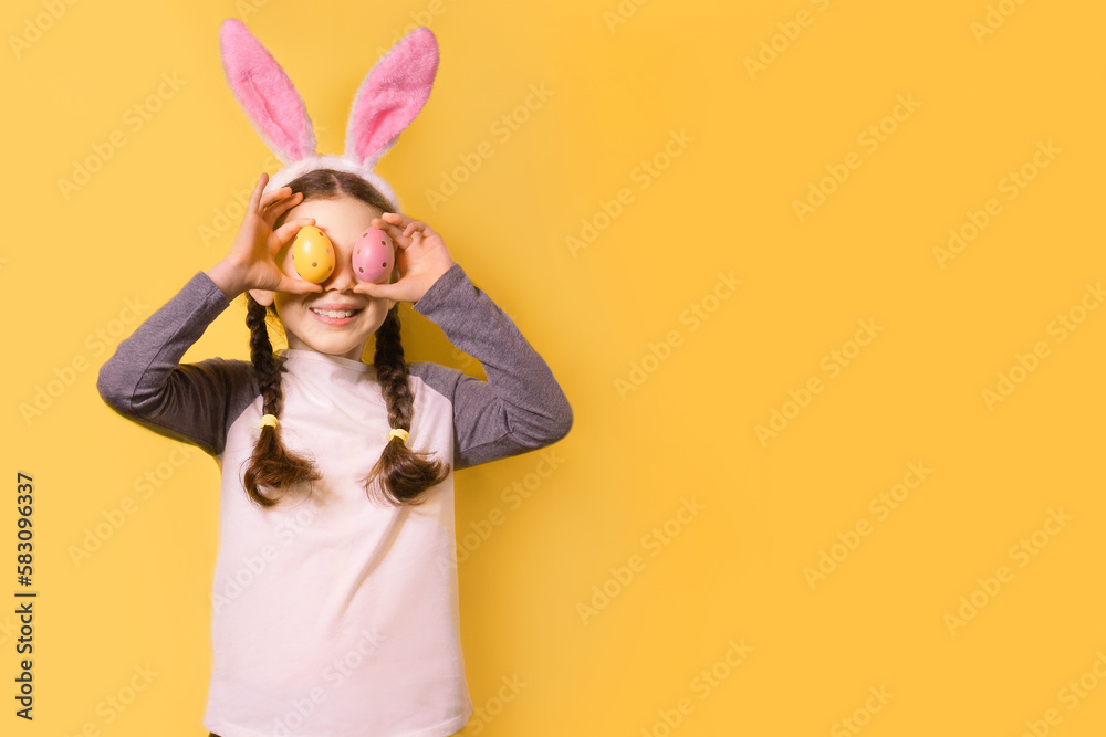 Funny happy girl with Easter bunny ears and colorful Easter eggs on a yellow background. Happy Easter! Copy space.