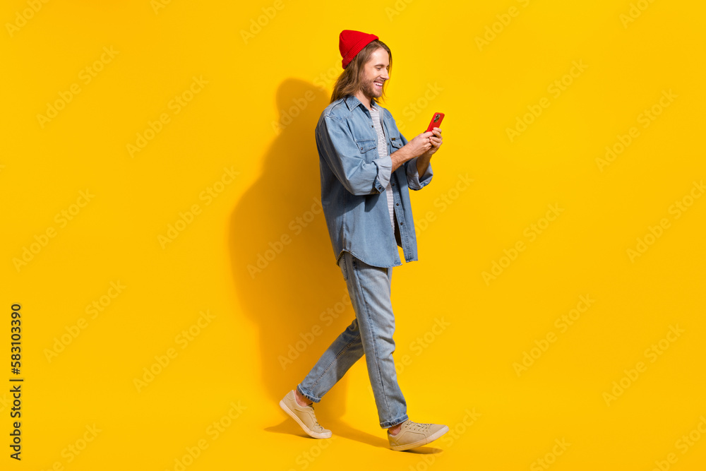 Naklejka premium Full size photo of positive man wear denim jacket jeans trousers hold smartphone walk empty space isolated on yellow color background