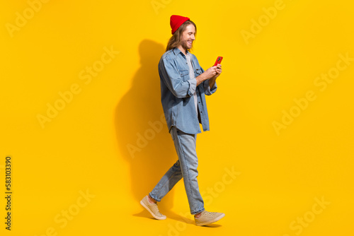 Full size photo of positive man wear denim jacket jeans trousers hold smartphone walk empty space isolated on yellow color background © deagreez