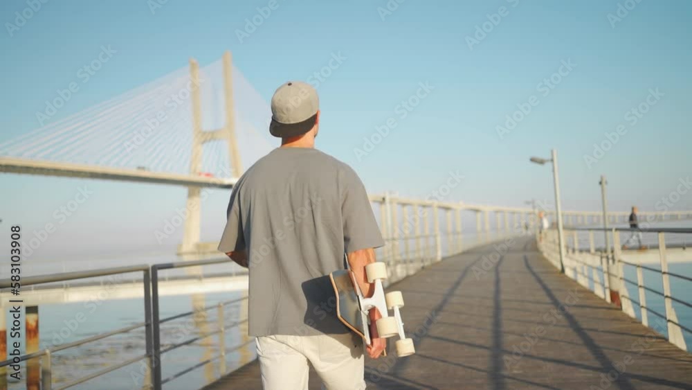 Man walk with skateboard on bridge summer day near ocean. Adult male person skateboarder enjoy after riding on skate. Millenial hipster with longboard 