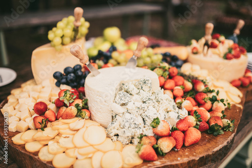 The board is set up on a long buffet table. The cheeses are arranged alongside sliced cured meats, fresh grapes, and berries, and the warm glow of the overhead lights adds to the overall ambiance 
