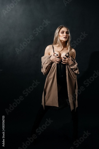 A girl in black underwear and a raincoat clutches her chest standing against a black background