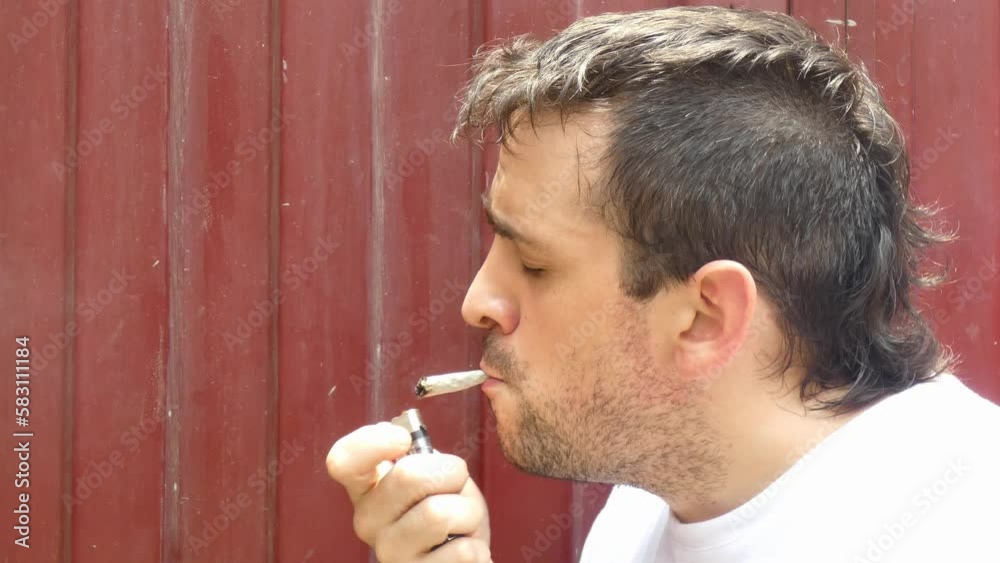 A person lighting a marijuana cigarette, or a cannabis joint in front of a red wall. The man who has a mohawk hairstyle lights the joint with one hand and then exhales the smoke.