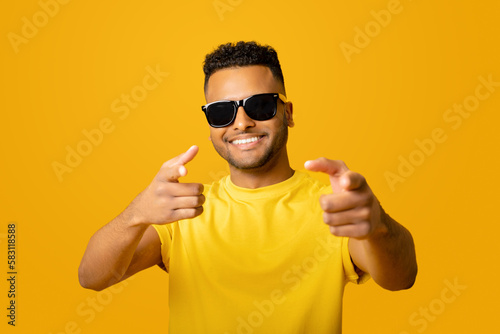 Hey you. Excited positive young arab man in sunglasses pointing fingers at the camera, choosing you, handsome indian guy looking at the camera and laughing, isolated on yellow background © Vadim Pastuh