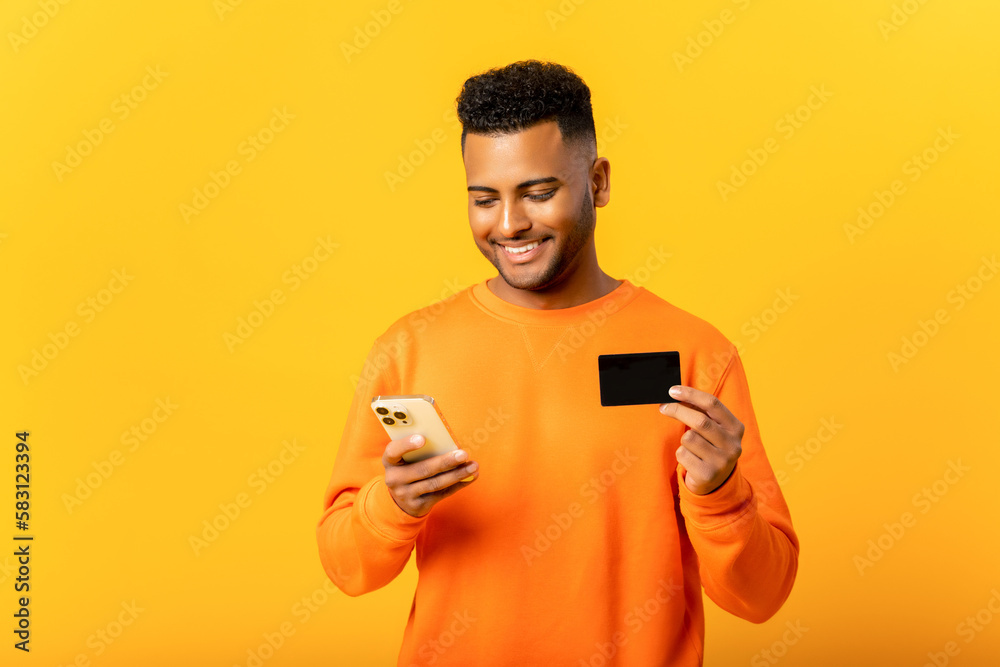 Carefree happy Indian man holding credit card and smartphone, looking at the smartphone screen isolated on yellow background, arab guy making order online, e-banking concept Carefree happy Indian man holding credit card and smartphone, looking at the smartphone screen isolated on yellow background, arab guy making order online, e-banking concept