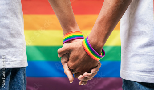 Fotografia Male couple wearing gay pride rainbow awareness wristbands holding hands on rainbow flag background