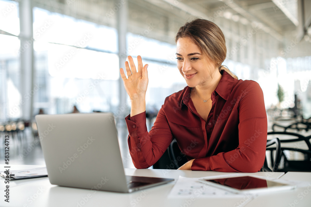Positive successful caucasian woman, company employee, manager, sitting at a work desk in the office, talking on online video conference with colleagues, making a welcome gesture, smiling Positive successful caucasian woman, company employee, manager, sitting at a work desk in the office, talking on online video conference with colleagues, making a welcome gesture, smiling