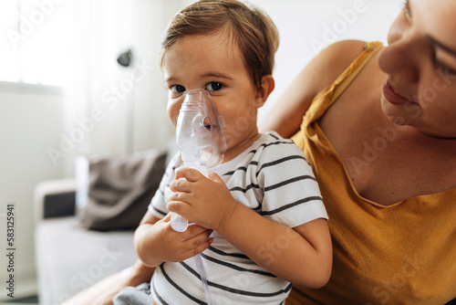 Portrait of a baby boy inhaling medicine in nebulizer with his mother at home.