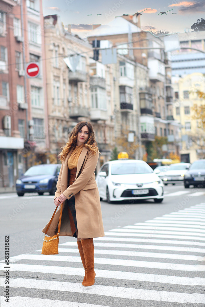 A modern brunette girl in a brown coat and high boots and a beige handbag walking across the road. Walk. Autumn. urban concept