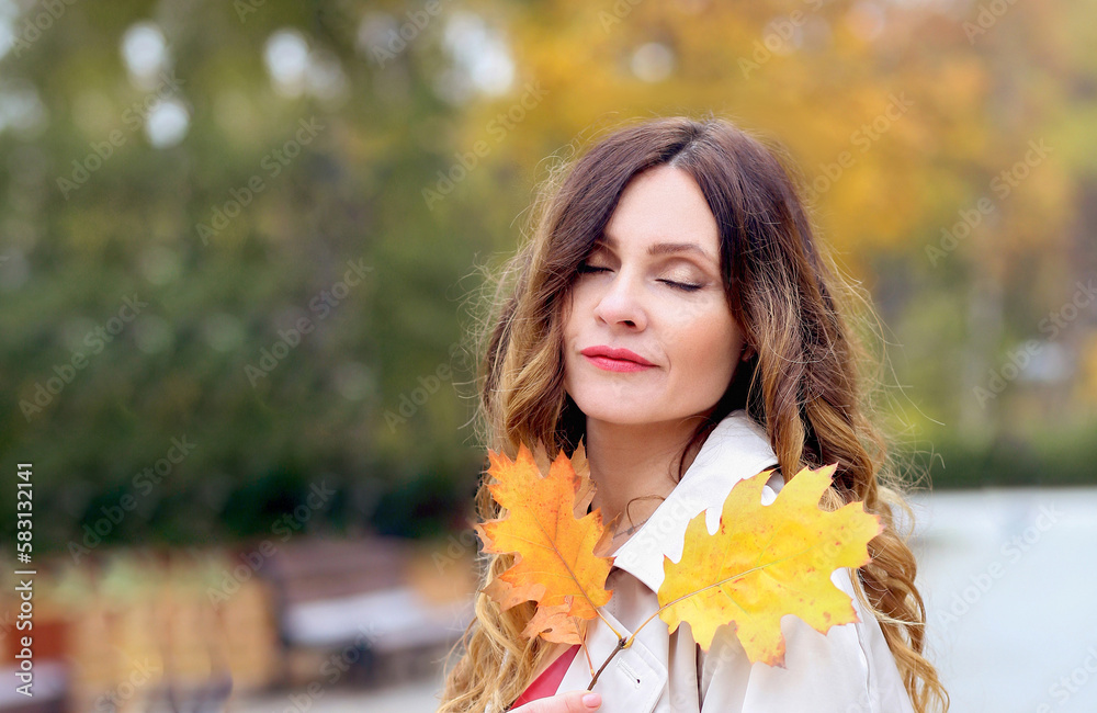 Banner with a close-up sensitive girl in a beige raincoat with yellow leaves close to her face on the background of a city park. Autumn concept. Copy space on the left