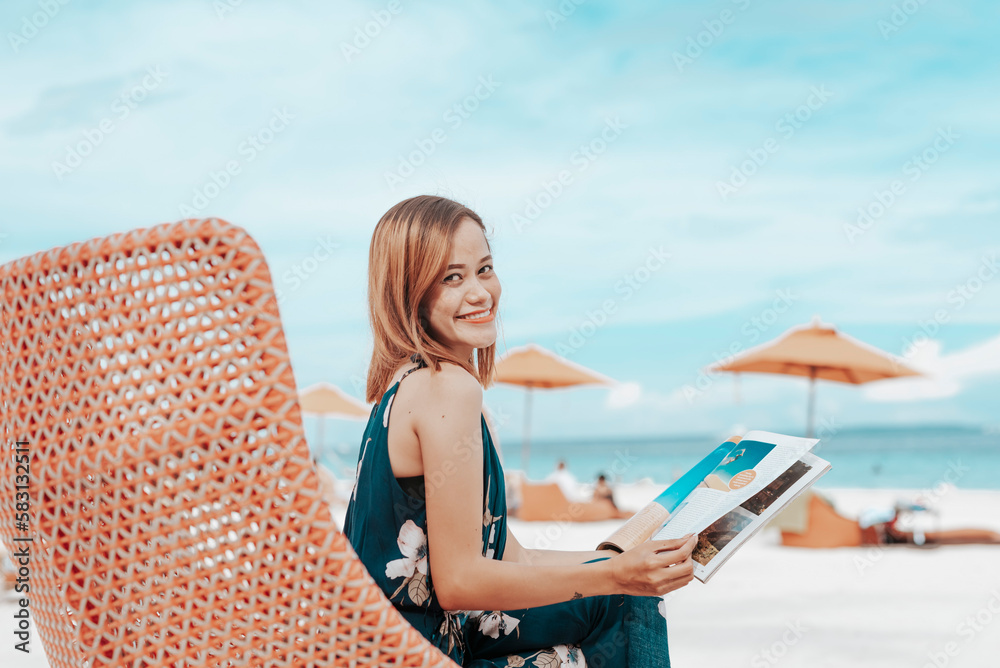 A young lady in a blue floral dress reading a lifestyle magazine while sitting on a comfy lounge chair near the beach. During her vacation at a tropical resort.
