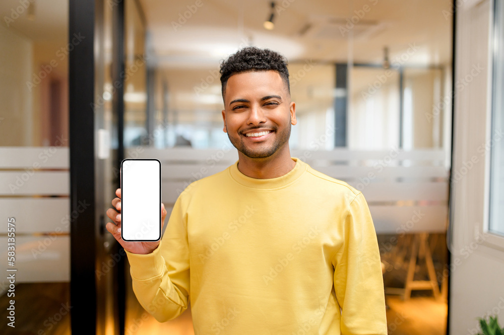 Serene smiling Indian man holding and showing smartphne with empty phone screen, guy presenting best deal, advertising new mobile app standing in modern office Serene smiling Indian man holding and showing smartphne with empty phone screen, guy presenting best deal, advertising new mobile app standing in modern office
