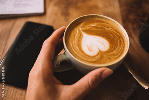 Soft focus effect. Cropped image of female holding ceramic coffee cup with aroma traditional hot Italian beverage, top view of late art on top of cappuccino foam for morning drink and awaken