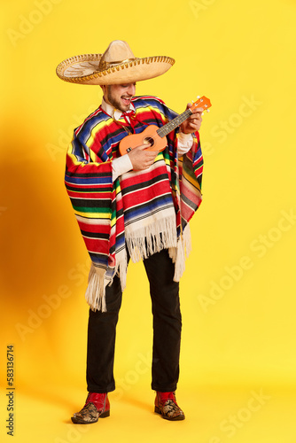 Quadro su tela Portrait of young handsome man in colorful clothes, poncho and sombrero playing guitar, posing against yellow studio background