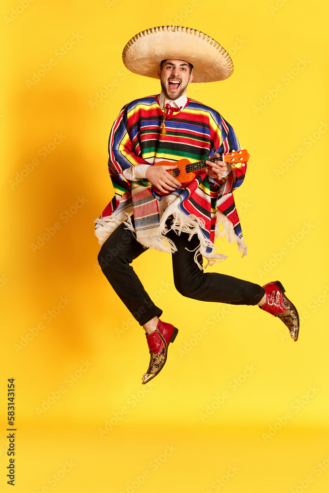 Young emotional man in colorful poncho and sombrero posing, playing guitar and jumping against yellow studio background. Concept of mexican traditions, fun, celebration, festival, emotions