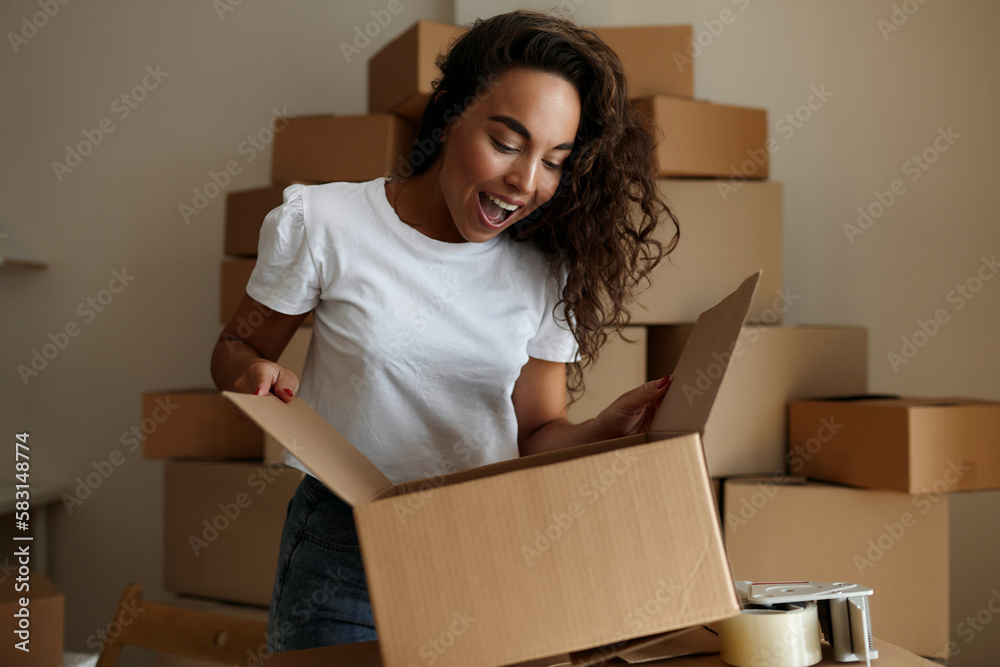 Pretty young woman in casual clothes stands at home with a postal parcel, unpacks a cardboard box, looking excited. Home delivery, shopping and online shopping concept Pretty young woman in casual clothes stands at home with a postal parcel, unpacks a cardboard box, looking excited. Home delivery, shopping and online shopping concept