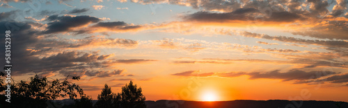 A summer sunset in yellow and orange colours. The sun's rays from below illuminate the dark clouds. Silhouettes of trees in the foreground and the silhouette of a mountain range in the background.