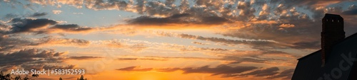 A summer sunset in yellow and orange colours. The sun's rays from below illuminate the dark clouds. In the foreground is a chimney on the roof of a house. Panoramic photo. © VeNN