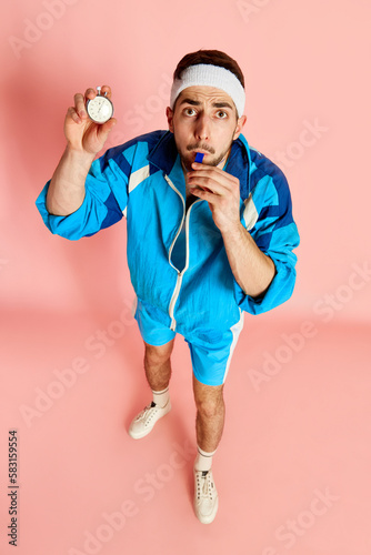 Top view image of fitness trainer, coach in blue uniform posing with whistle and taimer against pink studio background. Concept of sportive lifestyle, emotions, facial expression. Ad photo
