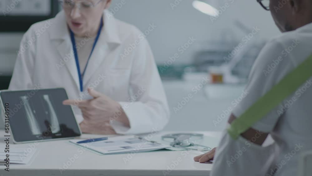 Racking focus of female practitioner showing x-ray image on tablet when consulting African American man with broken arm in cast sitting by desk at the doctors office Racking focus of female practitioner showing x-ray image on tablet when consulting African American man with broken arm in cast sitting by desk at the doctors office