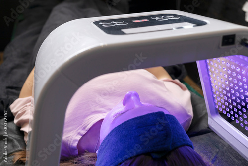 Young woman gets skin rejuvenation light treatment, lying on couch under blue led mask in spa salon. Facial skin therapy, care. Beauty face procedure. Horizontal plane, closeup.