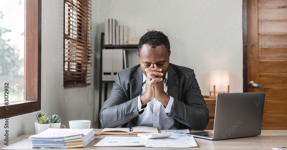 Fototapeta premium Stressed business man sitting at office workplace. Tired and overworked black man. Young african american exhausted men in stress working on laptop computer