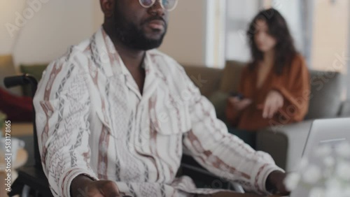 Loose close-up of black man in wheelchair opening laptop, putting headphones on, starting listening to music, with young dark-haird Caucasian woman sitting on couch holding smartphone, in background photo