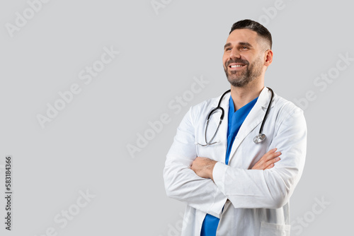 Confident male doctor in uniform posing with folded arms over grey studio background, looking at free space