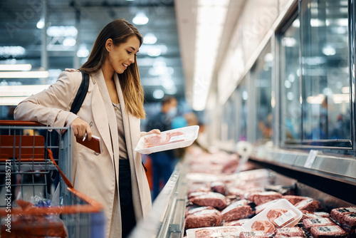 Happy woman buys meat at refrigerated section in supermarket.