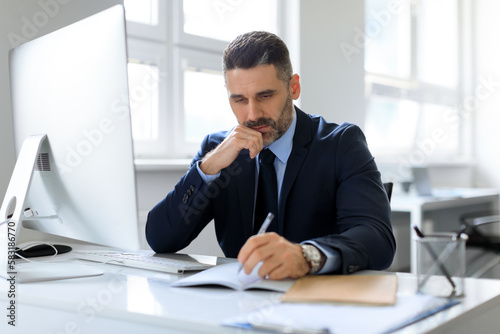 Focused caucasian middle aged businessman writing in notepad, planning working schedule, sitting at desk in office