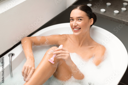 Attractive Lady Shaving Legs Smiling To Camera In Modern Bathroom