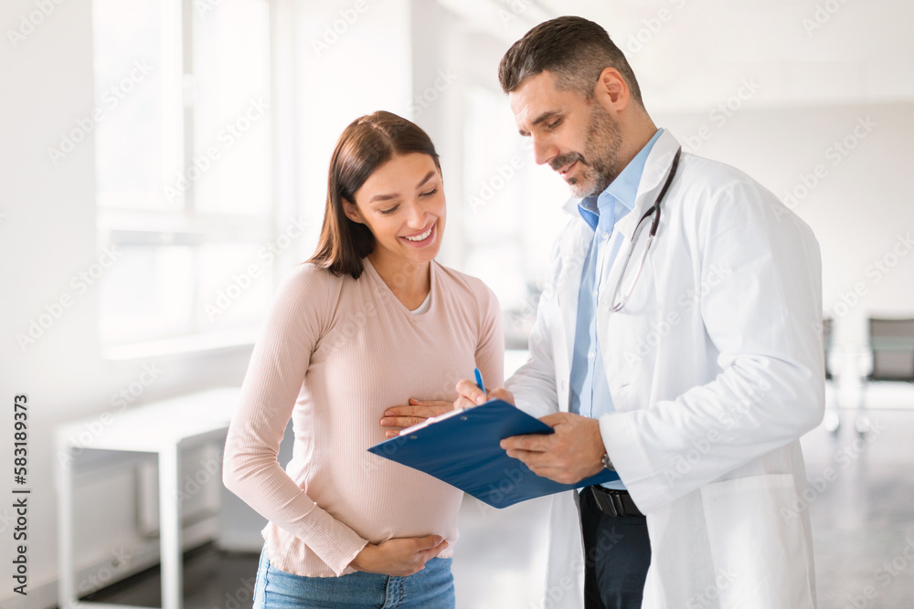 Male doctor showing clipboard with medical test results to happy pregnant lady during meeting in clinic Male doctor showing clipboard with medical test results to happy pregnant lady during meeting in clinic
