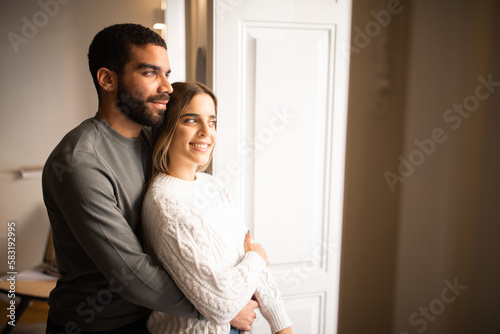 Happy multiethnic millennial couple hugging, look out the window at copy space, enjoy spare time together