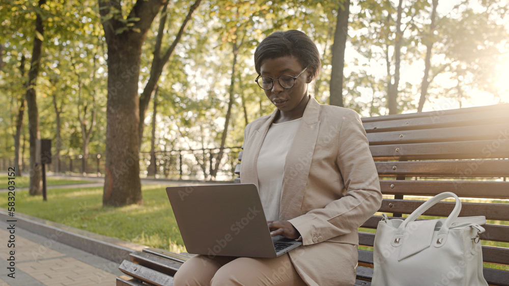 African American woman working on laptop, sitting on bench in summer park African American woman working on laptop, sitting on bench in summer park