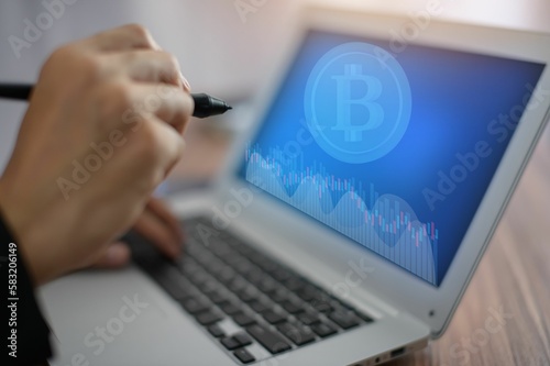 Hand holding a pen while typing in a laptop with the bitcoin symbol on the screen, close-up