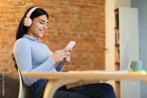 Quadro su tela Happy indian lady using cellphone, wearing wireless headphones and smiling, sitt