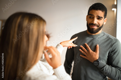 Married international millennial couple quarreling, wife pointing finger at husband in living room interior