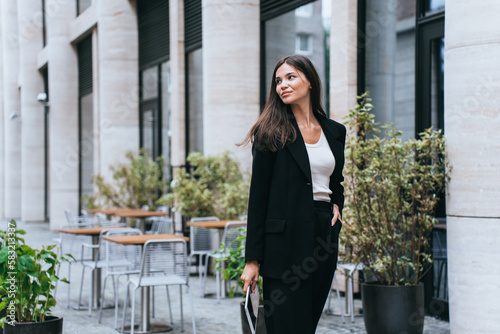 Pensive Asian brunette young businesswoman in black suit against building holds laptop looks at camera. Attractive female entrepreneur planning day. Purposeful student girl preparing for exam.